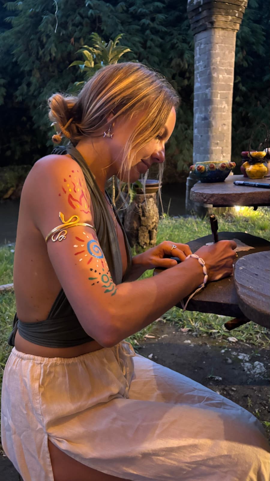 Lina at dusk with painted sacred symbols on her arm, preparing quietly at a wooden table in a garden.