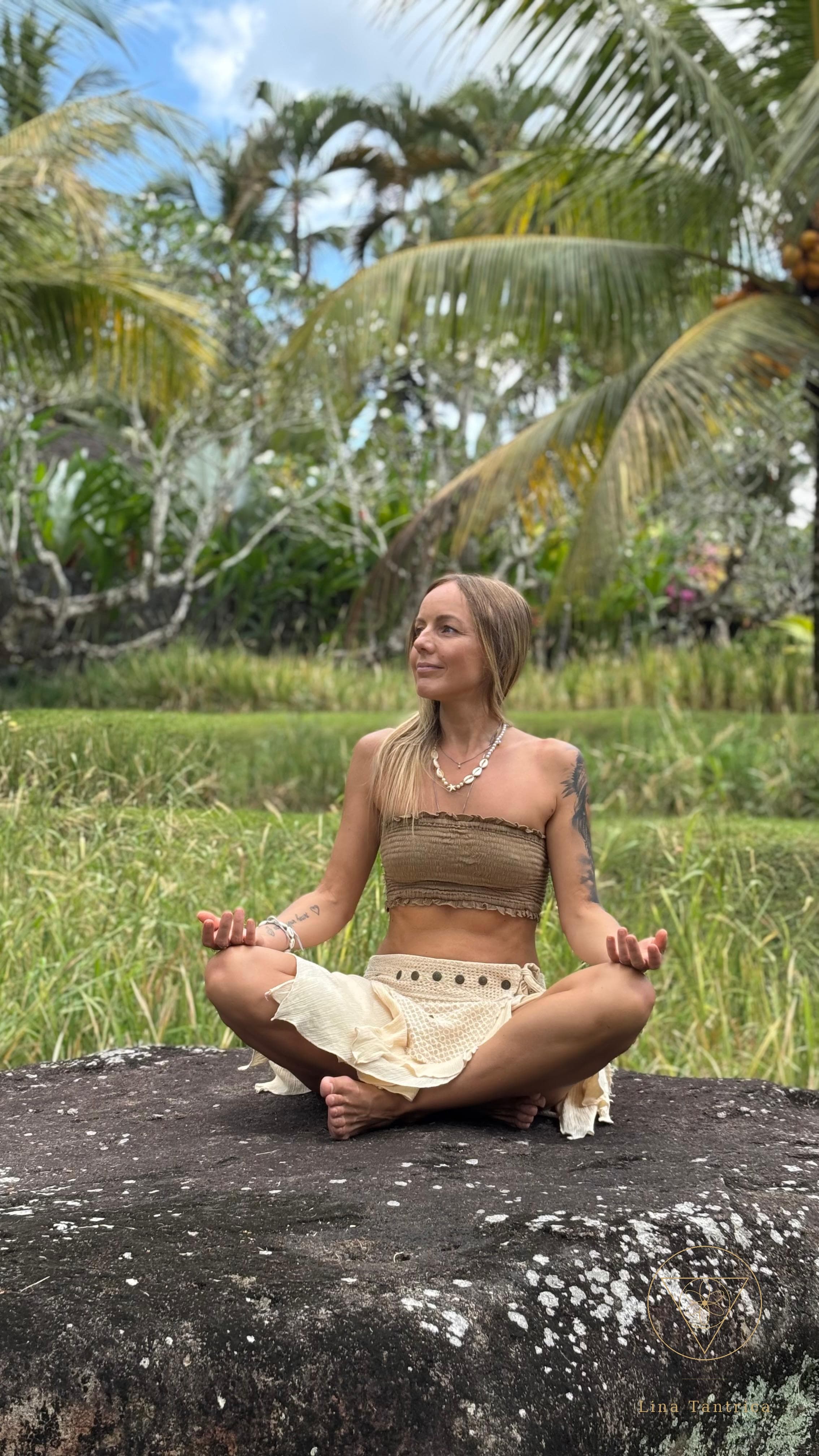 Lina seated cross-legged in meditation on a large stone, surrounded by tropical greenery and palms.