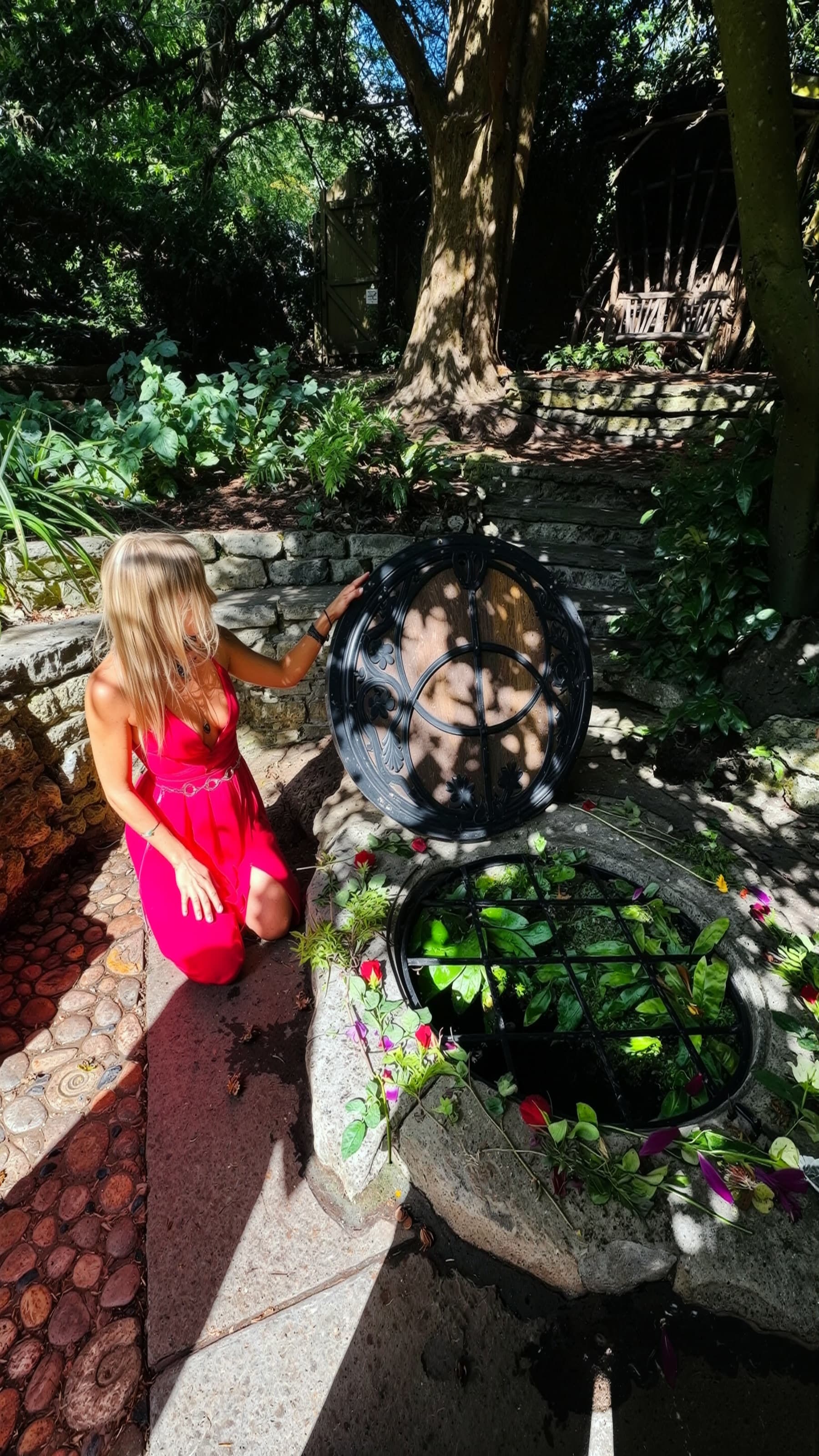 Lina kneeling beside the open lid of the Chalice Well, its sacred waters dressed with roses and flowers in a green garden.