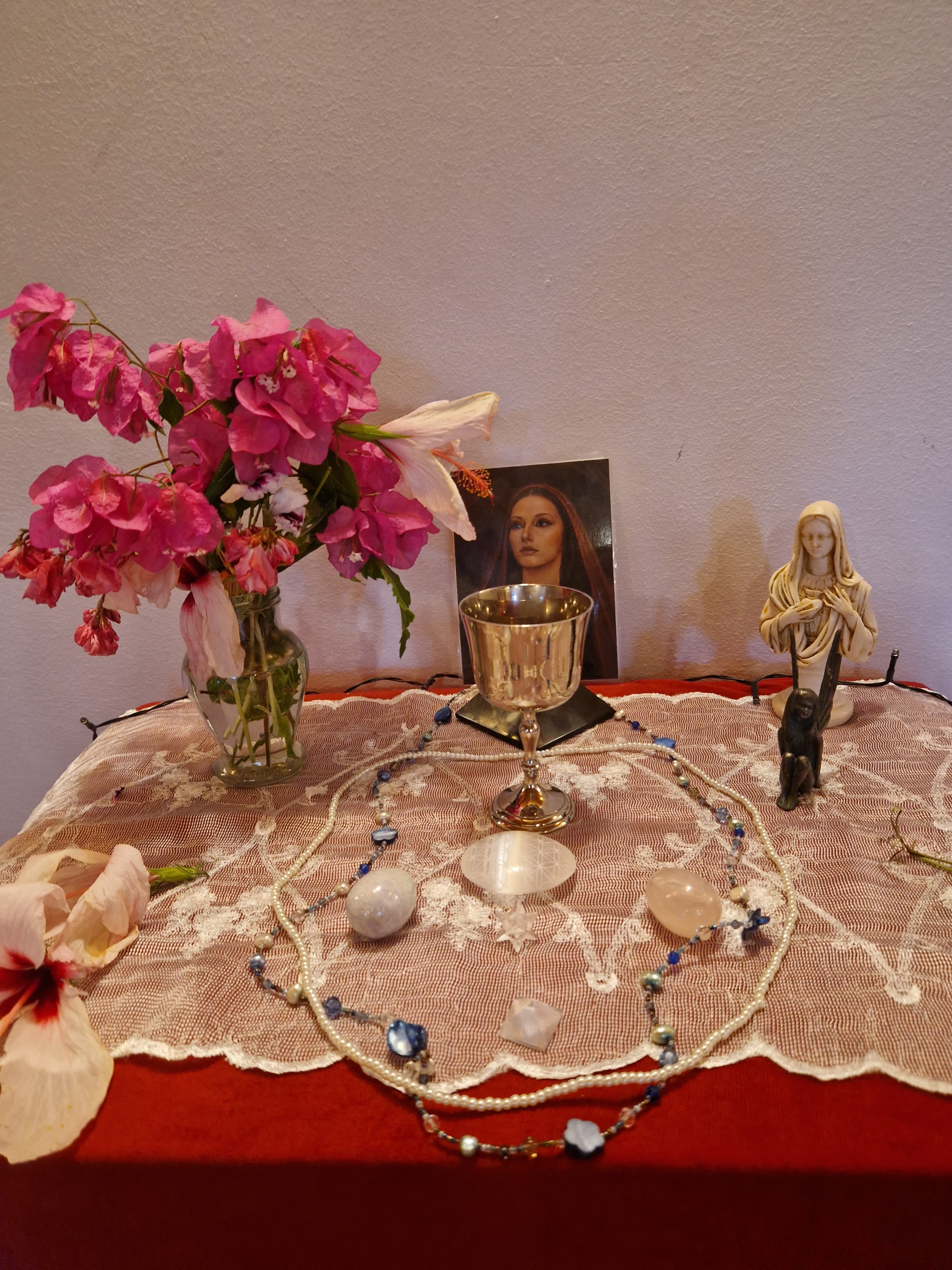 A devotional altar with a silver chalice, a rose-veiled image of the sacred feminine, a Madonna statue, crystals, pearls, and bougainvillea on lace.