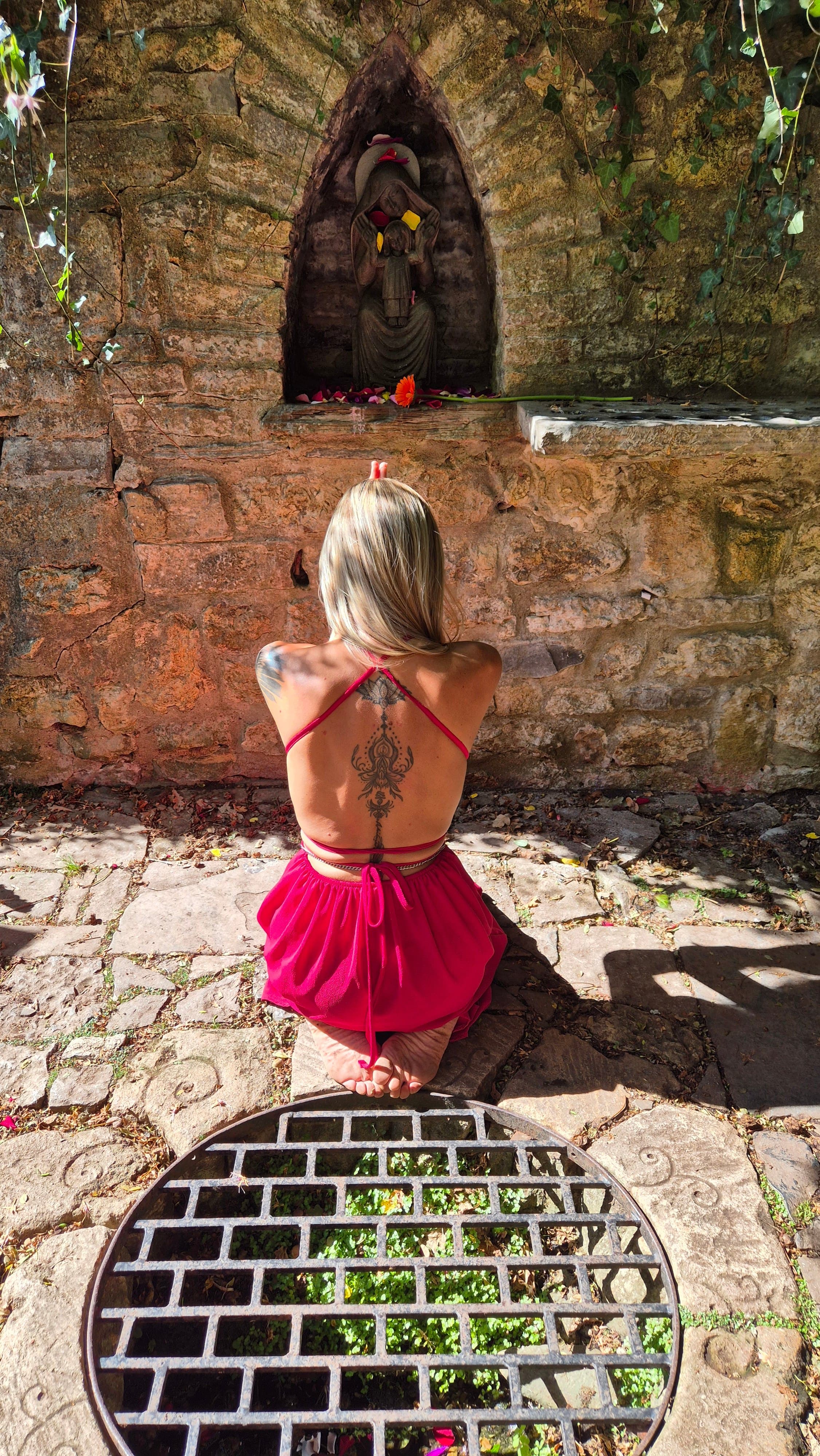 Lina kneeling in prayer before a weathered stone shrine niche scattered with rose petals as offerings.