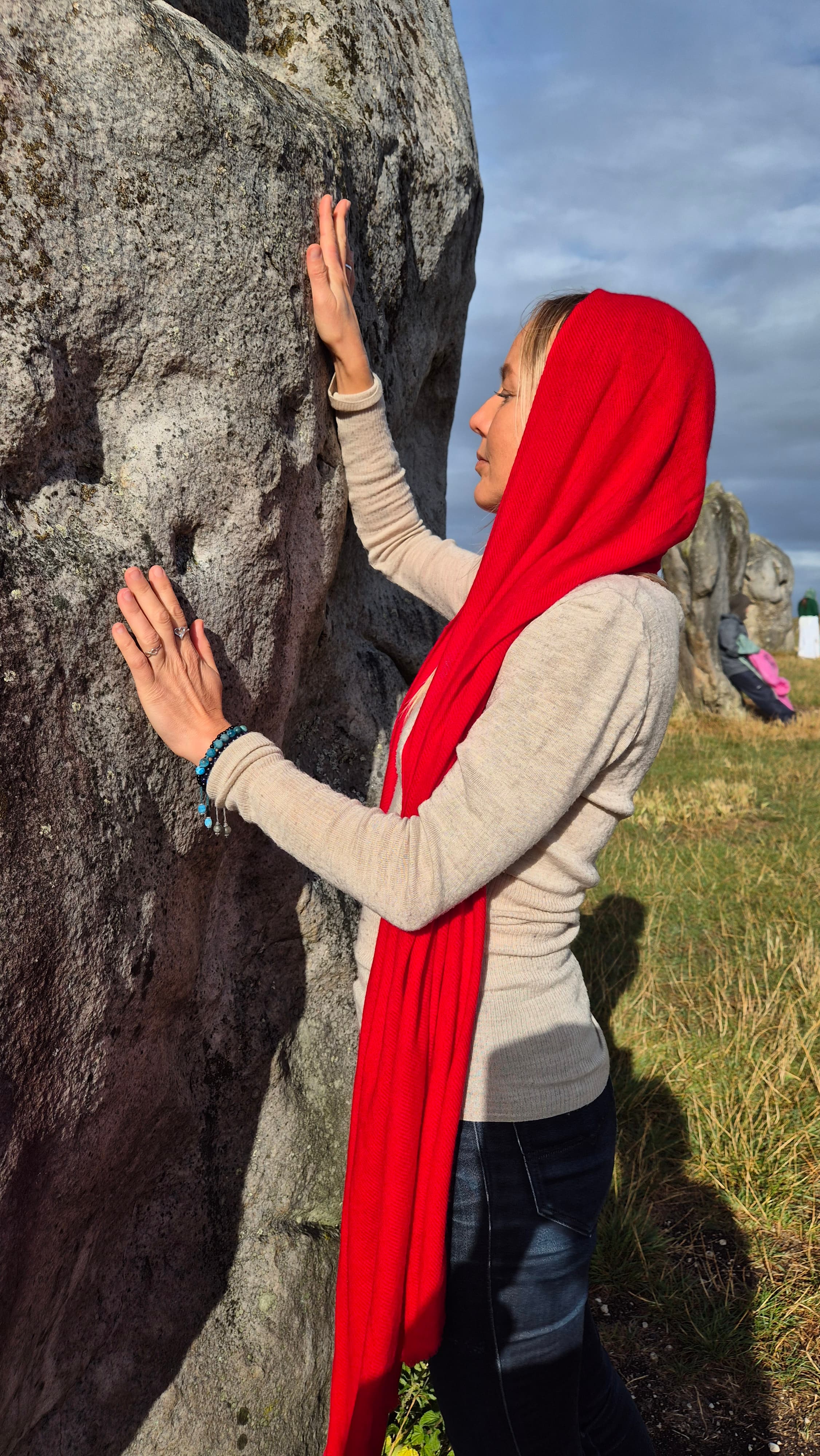 Lina in a red veil resting her hands in devotion against an ancient standing stone at a stone circle.