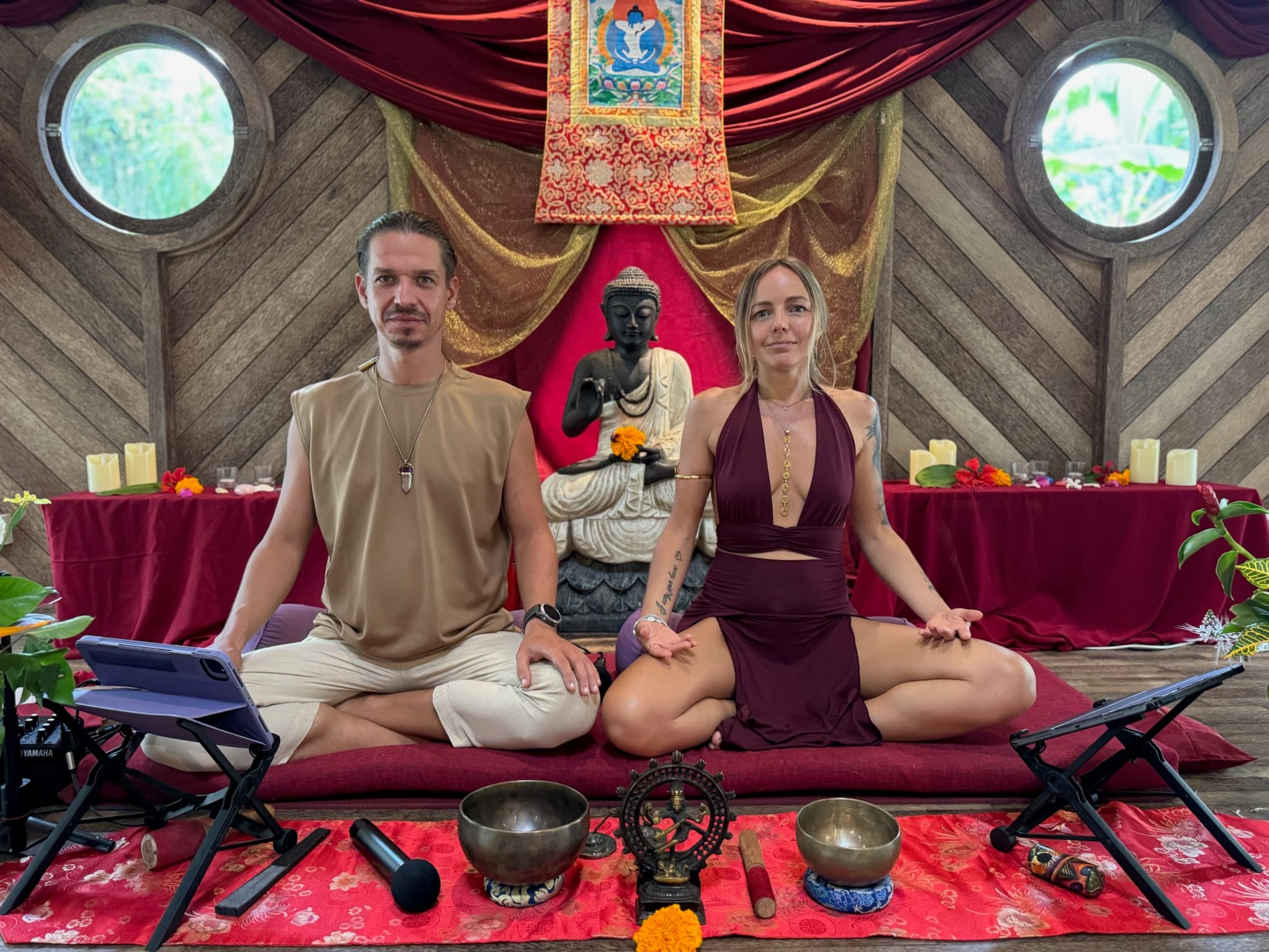 Lina and Ritam seated together in meditation posture on a burgundy mat in the Bali temple, a stone Buddha, thangka, and singing bowls around them.