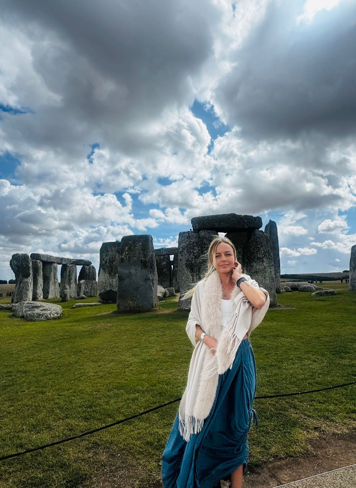 Lina in a cream shawl and teal skirt standing before the ancient stones of Stonehenge under a dramatic cloudy sky.